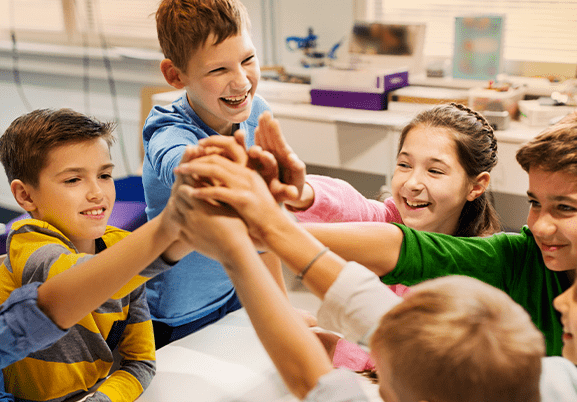 5 crianças, entre 8 e 10 anos, em ambiente escolar, juntando as mãos em comemoração no centro da mesa