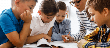 Ensino Fundamental – 4 crianças entre 7 e 9 anos em ambiente escolar junto com uma professora conferindo material educacional