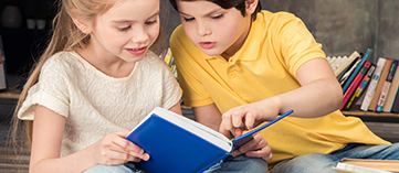 Educação Infantil- menina loira e menino moreno, entre 4 e 6 anos, sentados lendo um livro de capa azul.